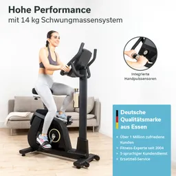 Fitnessbike mit 14 kg Schwungmasse. Person sitzt darauf, hält Lenker, graue Leggings, neben einem Sofa. Handpulssensoren sichtbar.