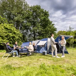 Gruppe von fünf Personen sitzt und steht um ein Zelt auf einer Wiese, umgeben von Bäumen. Tisch mit Getränken und Essen.