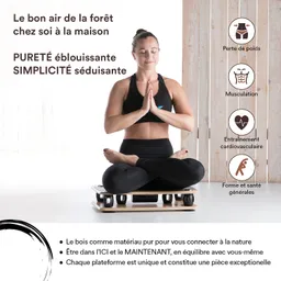 Eine Person sitzt im Schneidersitz auf einer Plattform, hält die Hände vor der Brust in einer Meditationshaltung. Die Person trägt sportliche Kleidung. Im Hintergrund sind helles Holz und Fenster zu sehen.