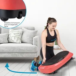 Fitnessgerät in Rot auf einem hellen Boden, neben einer grauen Couch mit Kissen. Eine Person kniet und hebt das Gerät an.
