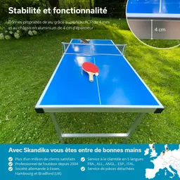 Tischtennistisch mit blauer Oberfläche und Netz, Schläger auf der Platte, im Freien auf grünem Gras.