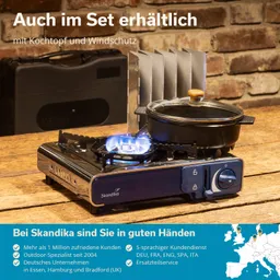 Kocher auf Holztisch mit flammendem Gas, Kochtopf mit Glasdeckel und Windschutz aus Metall, im Hintergrund eine schwarze Tasche.