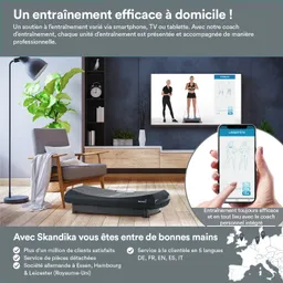 Ein Wohnraum mit einem grauen Fußtrainer im Vordergrund, einem Fernseher an der Wand mit Trainingsanleitung und einem Smartphone in der Hand einer Person. Eine Pflanze und eine Lampe sind ebenfalls sichtbar.