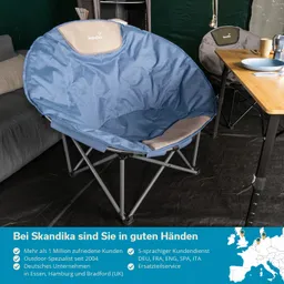 Ein blauer, runder Klappstuhl mit grauem Polster auf einem dunklen Boden. Im Hintergrund ein Tisch mit Geschirr.