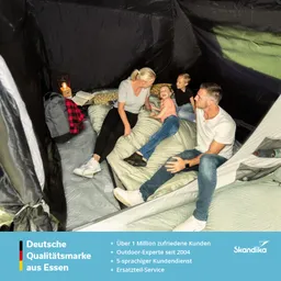 Vier Personen sitzen in einem Zelt auf Schlafsäcken. Eine Person hält ein Buch, während die anderen lächeln und spielen. Links steht ein Rucksack und eine lampe.