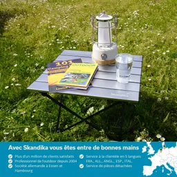 Klappbarer Tisch aus Aluminium mit zwei Campingbüchern, einer Lampe und einem Glas Wasser auf einer Wiese.