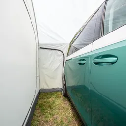 Ein grünes Auto steht neben einer weißen Zeltwand. Der Zelteingang ist geöffnet und führt nach außen. Grass ist sichtbar.