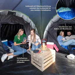Zwei Personen sitzen im Zelt auf dem Boden. In der Mitte steht eine Holzkiste mit verschiedenen Gegenständen. Im Hintergrund sind Schlafkabinen sichtbar.