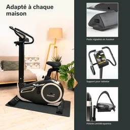 Crosstrainer auf einem Teppich in einem Wohnraum, mit einem Sofa, Pflanze und Stehlampe im Hintergrund.