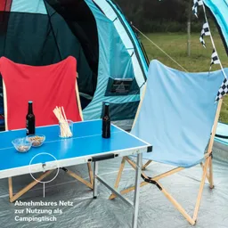 Zwei Stühle, rot und blau, stehen an einem blauen Tisch. Darüber sind zwei Flaschen und Snacks. Im Hintergrund ein Zelt und Wiese.