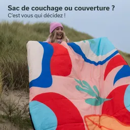 Eine Person hält einen farbenfrohen Schlafsack oder eine Decke in einer Landschaft mit Gras und Sand.
