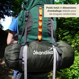 Rucksack in grün mit orangefarbenen Akzenten, auf dem Rücken einer Person. Darunter eine schwarze, zusammengerollte Ausrüstung. Hintergrund mit Bäumen.