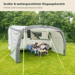 Stellplatz mit Wohnwagen und einem überdachten Bereich. Zwei Personen sitzen auf Campingstühlen und trinken Kaffee.