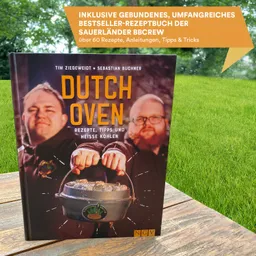 Ein Kochbuch mit dem Titel "Dutch Oven" liegt auf einer Holzoberfläche. Im Hintergrund ist grünes Gras sichtbar.
