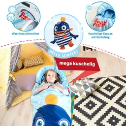 Kind liegt in einem Schlafsack mit Tiermotiv auf einem Teppich. Im Hintergrund steht ein Zelt mit Kissen. Farbige Muster sind sichtbar.