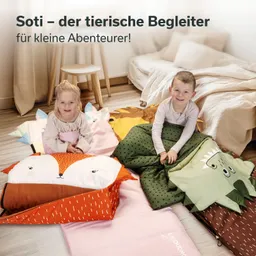 Zwei Kinder sitzen auf bunten Kissen und Decken im Raum. Eines der Kissen zeigt einen Fuchs, ein anderes ein Tier mit Augen. Im Hintergrund sind Möbel sichtbar.