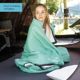 Eine Person sitzt auf einer Matratze in einem Zelt, eingehüllt in einen mintgrünen Schlafsack. Im Hintergrund sind Zelte und ein Tisch sichtbar.