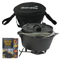 Dutch Oven aus Gusseisen mit Deckel und Füßen, daneben eine schwarze Tragetasche und ein Rezeptbuch mit verschiedenen Gerichten.