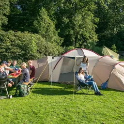 Eine Gruppe von Personen sitzt an einem Tisch mit Getränken und Essen. Im Hintergrund steht ein großes Zelt. Zwei Personen sind vor dem Zelt, eine sitzt auf einem Stuhl mit einem Buch. Umgebende Bäume sind sichtbar.