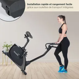 Fahrrad-Ergometer mit Transportrollen und einer Person, die es hält. Im Hintergrund eine Pflanze und ein Holzboden.