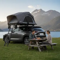 Zwei Personen sitzen an einem Holztisch mit Essen und Getränken. Im Hintergrund steht ein Auto mit einem Dachzelt, vor einer Berglandschaft.