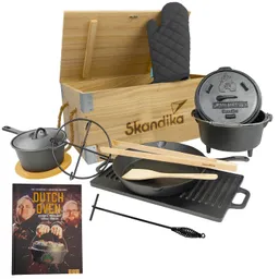 Holzkiste mit "Skandika"-Logo, gefüllt mit einem Dutch Oven, Pfanne, Kochtopf, Grillplatte, Kochutensilien und einem Kochbuch.