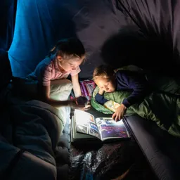 Zwei Kinder sitzen in einem Zelt. Eines hält eine Taschenlampe, während das andere ein Buch betrachtet. Decken sind um sie gewickelt.