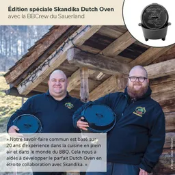 Zwei Personen halten Dutch Ovens vor einer rustikalen Holzstruktur. Die Landschaft im Hintergrund ist hügelig und bewaldet.