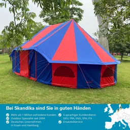 Ein großes, buntes Zelt mit blauen und roten Segmenten steht auf einer grünen Wiese, umgeben von Bäumen.