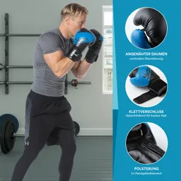 Mann in Sportkleidung trägt blaue und schwarze Boxhandschuhe. Er steht in einem Fitnessraum. Gewichte im Hintergrund sichtbar.