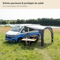 Ein blauer VW-Bus steht auf einer Wiese. Darunter ein Markisenbereich mit einem Tisch und zwei Stühlen.
