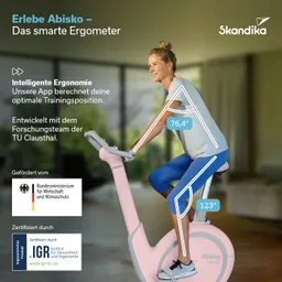 Ergometer in hellem Design mit einer Person, die darauf sitzt. Grüne Pflanzen im Hintergrund.