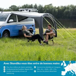 Ein blau-weißer Camper steht auf einer Wiese. Zwei Personen sitzen unter einem grauen Vorzelt, eine hat eine Tasse, die andere liest. Im Hintergrund ist ein Gewässer und Bäume zu sehen.