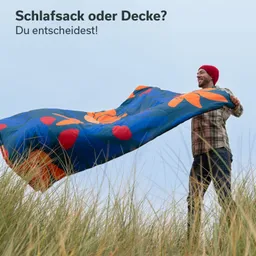 Person in kariertem Hemd und roter Mütze hält buntes Tuch in der Hand, umgeben von hohem Gras. Himmel ist bewölkt.