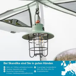 Grüne Campinglampe mit Schutzgitter hängt unter einem Zelt im Hintergrund Zeltmaterial und Teil einer Ausrüstung sichtbar.