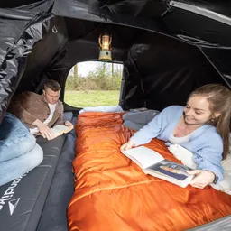 Zwei Personen lesen innerhalb eines Zeltes auf Schlafsäcken. Eine Person sitzt auf einem Luftmatratze, die andere liegt auf einem orangefarbenen Schlafsack. Eine Laterne hängt über ihnen. Im Hintergrund sind Bäume sichtbar.