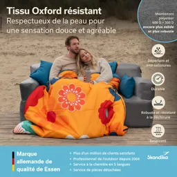 Ein Paar sitzt auf einem Sofa im Freien, eingekuschelt in eine bunte Decke mit Blumenmuster. Im Hintergrund ist ein Sandstrand zu sehen.