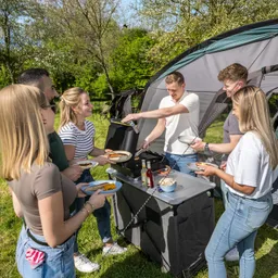 Grillstation im Freien mit sechs Personen, die Essen servieren und genießen. Zelt im Hintergrund, sonniger Tag.