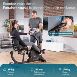 Eine Person sitzt auf einem Trainingsgerät, während eine andere Person Anweisungen gibt. Der Raum hat einen modernen Stil mit einem Sofa und einem TV im Hintergrund.