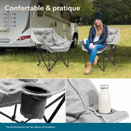 Zwei graue Faltstühle stehen auf einem Rasen neben einem Wohnmobil. Eine Person sitzt auf einem Stuhl. Auf dem anderen Stuhl ist ein Getränkehalter sichtbar, in dem eine Flasche steht.