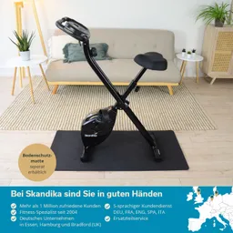 Widerstandstrainer mit schwarzem Rahmen und gepolsterter Sitzfläche, auf einer Bodenschutzmatte. Im Hintergrund Sofa und Pflanzen.