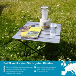 Ein klappbarer Tisch steht auf einer Wiese mit Blumen. Auf dem Tisch liegen zwei Campingbücher, eine weiße Laterne und ein Glas Wasser.