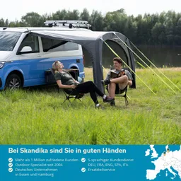 Zwei Personen sitzen auf Klappstühlen neben einem blauen Wohnmobil unter einem Gestell. Im Hintergrund gibt es Wasser und Bäume.