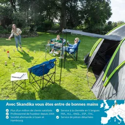 Drei Personen im Freien, eine wirft einen Gegenstand, zwei sitzen entspannt auf Campingstühlen. Zelt und Tisch mit Früchten sind sichtbar.