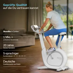 Stationäres Fahrrad mit einem Mann im kurzen Outfit. Display zeigt Trainingsdaten. Heller Raum mit Pflanzen im Hintergrund.