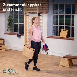 Eine Person trägt ein holzfarbenes, zusammenklappbares Fitnessgerät in einer Hand und elastische Bänder in der anderen Hand. Im Hintergrund sind eine Wand und Fenster sichtbar, sowie weitere Fitnessgeräte.