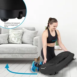 Eine Person in Sportkleidung hebt ein schwarzes Fitnessgerät. Im Hintergrund steht ein graues Sofa mit Kissen.
