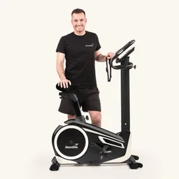 Ein Fitnessgerät, ein Ergometer, steht im Vordergrund. Daneben steht eine Person mit einem schwarzen T-Shirt und Shorts und lächelt.