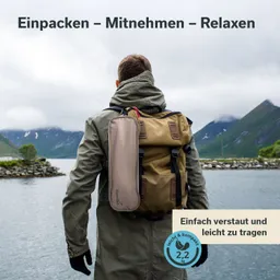 Person mit Rucksack steht am Wasser, umgeben von Bergen und bewölktem Himmel. Text: "Einpacken – Mitnehmen – Relaxen".