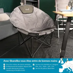 Grau gepolsterter Stuhl mit schwarzem Gestell auf schwarzem Boden, im Hintergrund weitere Möbel und grüne Wand.